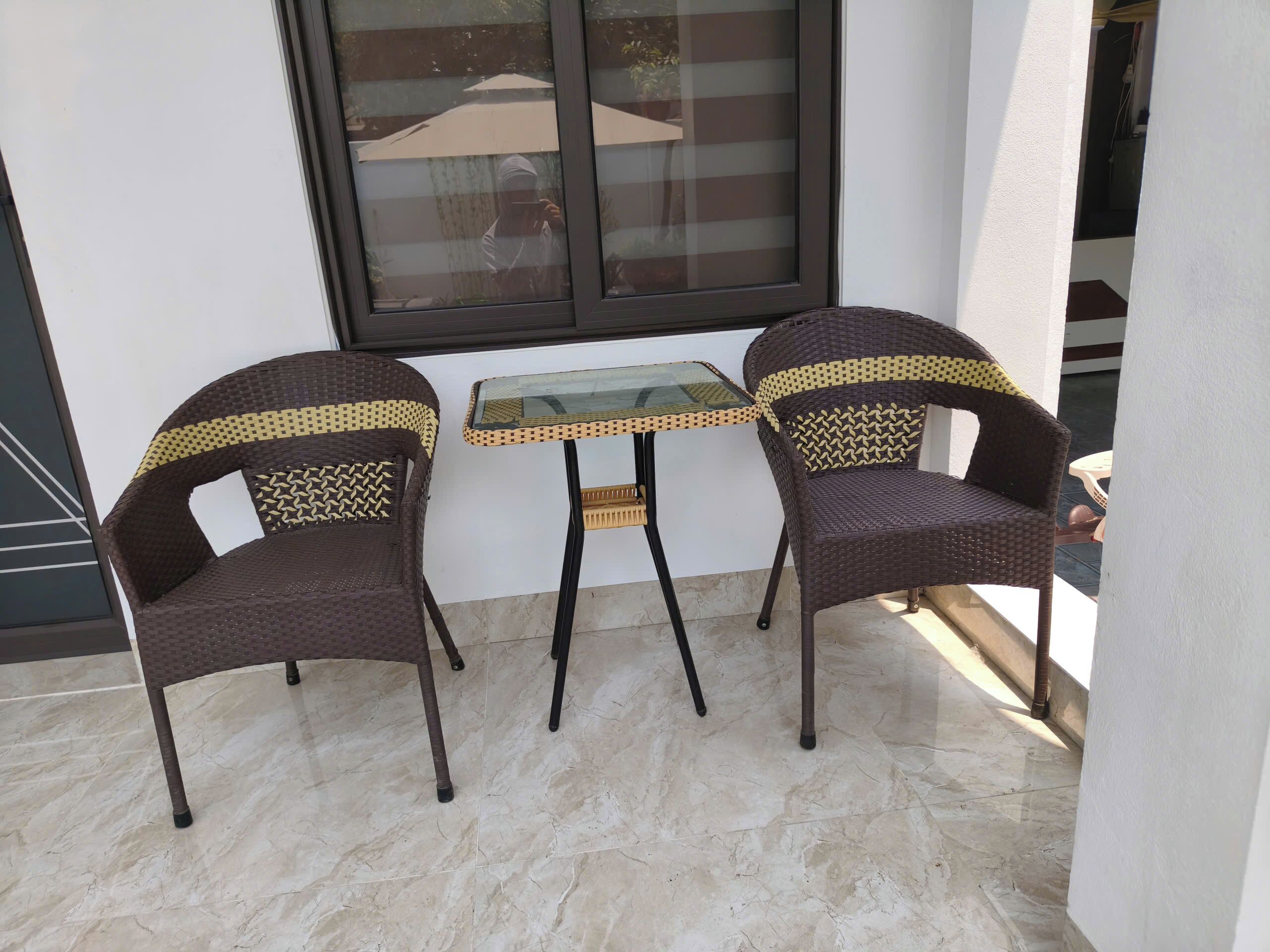 Duas cadeiras de vime com padrão trançado, uma mesa com tampo de vidro e uma janela com persianas.