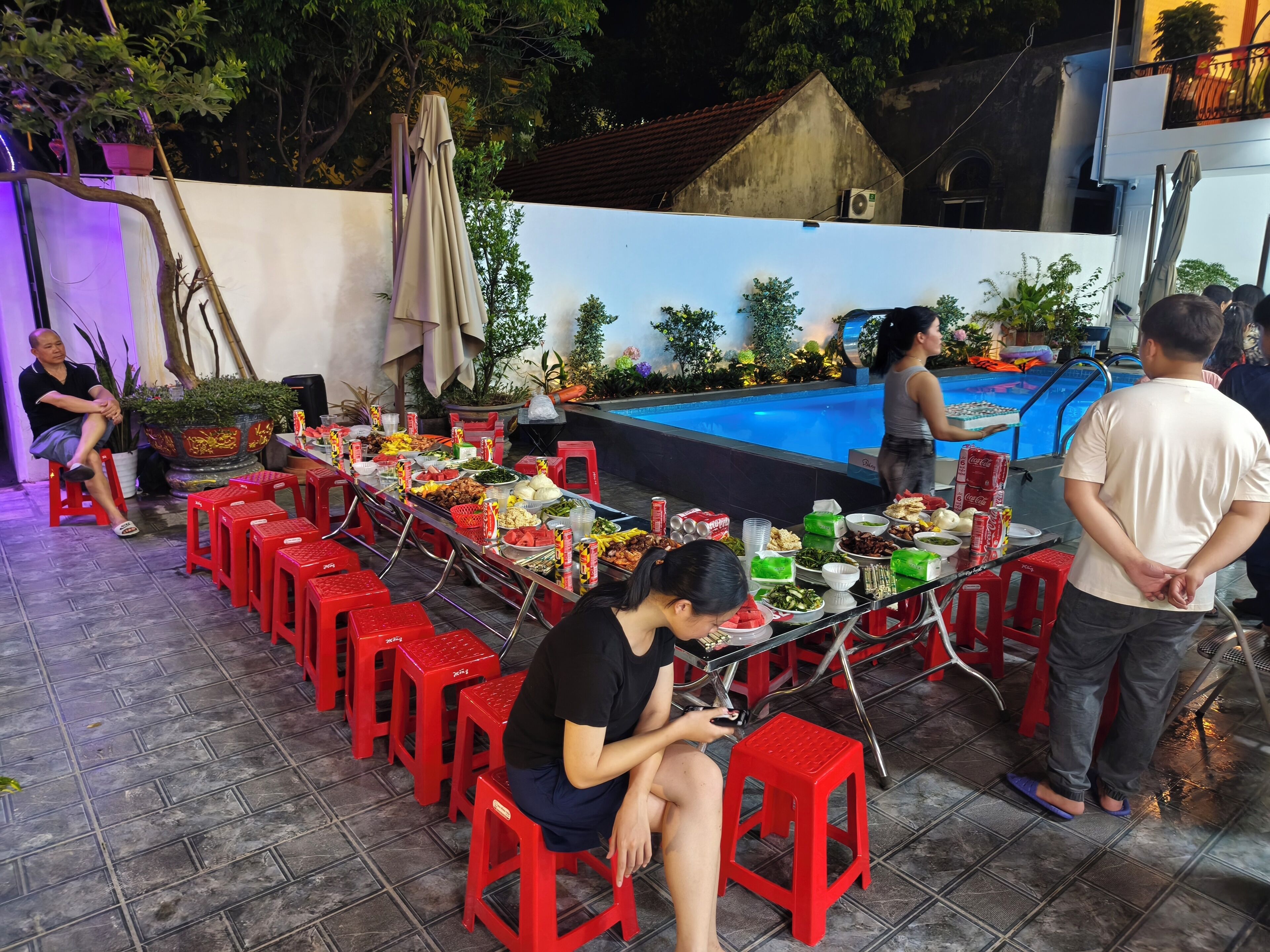 A dining area with tables set for a meal, surrounded by red stools, and a pool in the background.