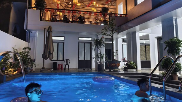 Piscina com duas pessoas, área externa coberta com plantas e varanda com guarda-corpo.