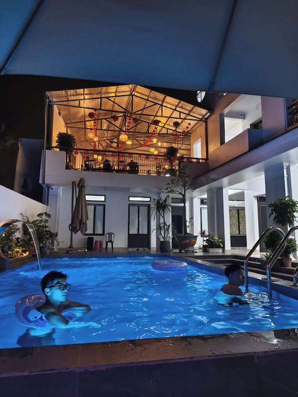 A swimming pool with two people, a covered outdoor area with plants, and a balcony with railings.