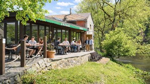 A restaurant with outdoor seating overlooking a river, stone walls, and greenery. - Hôtel La Ferme de Sy (Ferrières)