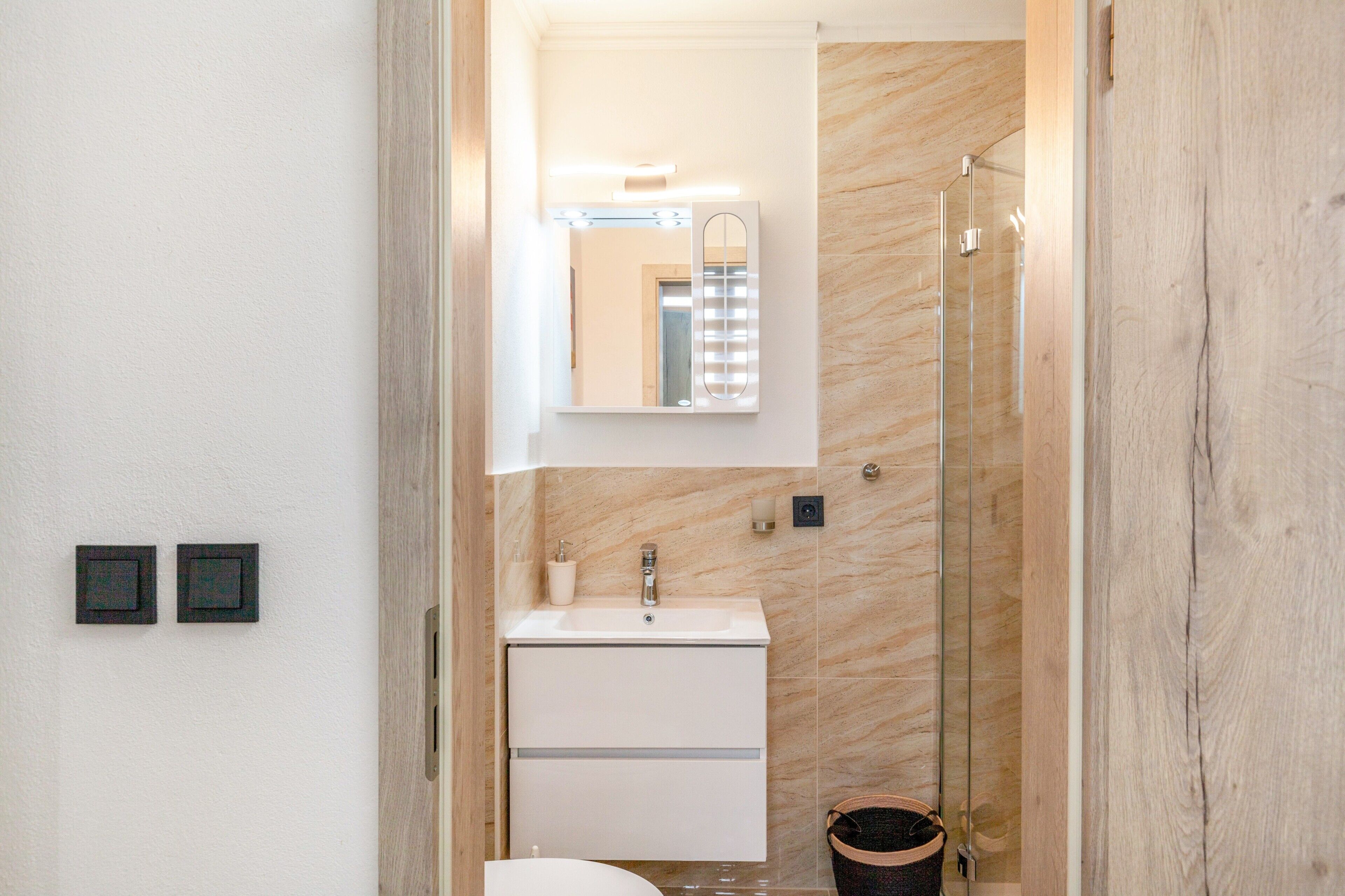 Une salle de bain avec une douche en verre, un lavabo blanc, un miroir et deux objets carrés noirs accrochés au mur.