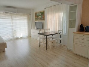 A room with a dining table, chairs, a TV, a cabinet, and a window with blinds. - Bright Seaside Apartment in Cala Millor | Beach Nearby (Son Servera)