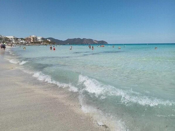 A beach with clear water, people swimming, and buildings in the background. - Bright Seaside Apartment in Cala Millor | Beach Nearby (Son Servera)