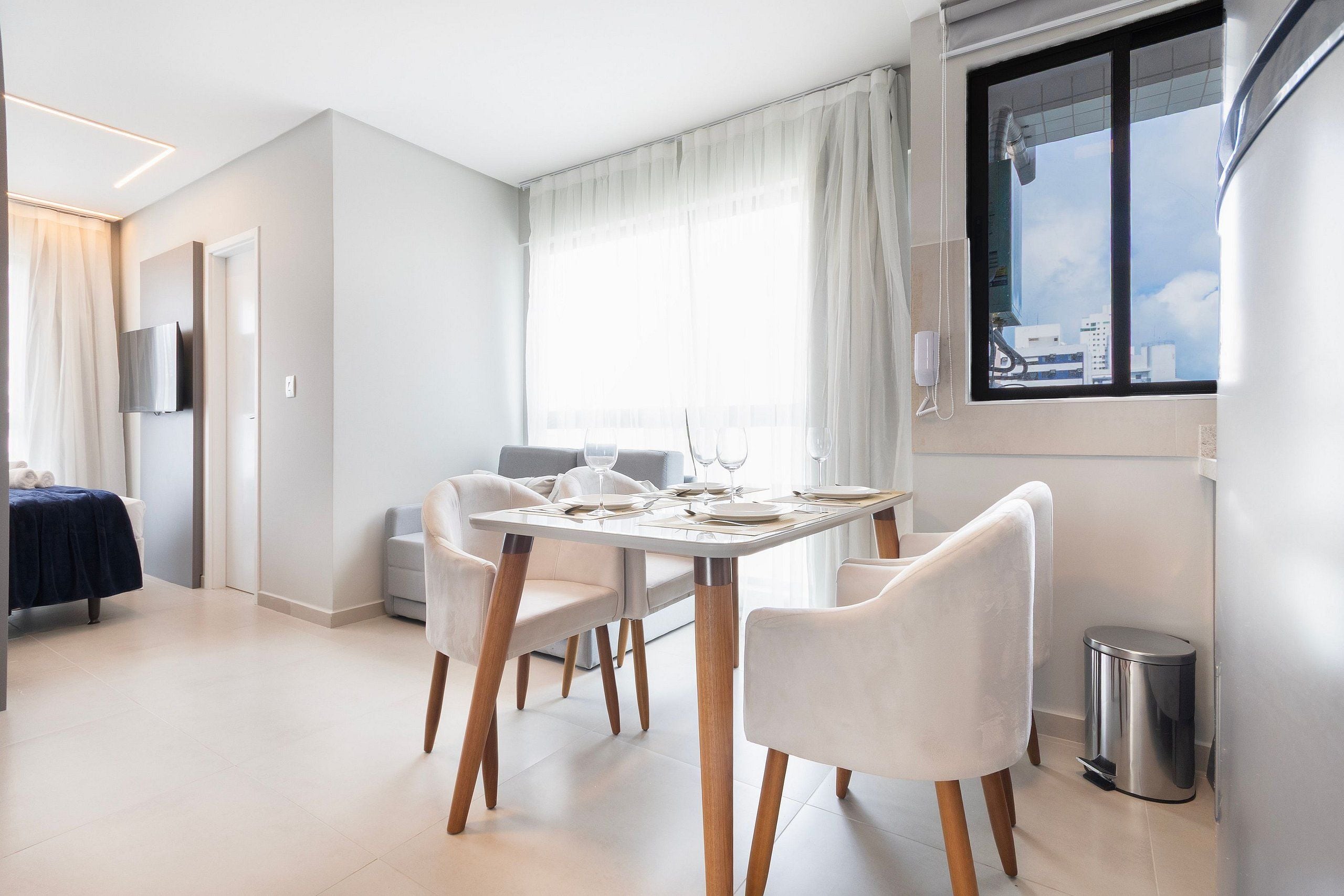 Un comedor moderno con una mesa dispuesta para cuatro personas, un sofá, un televisor y un ventanal con vistas a la ciudad.