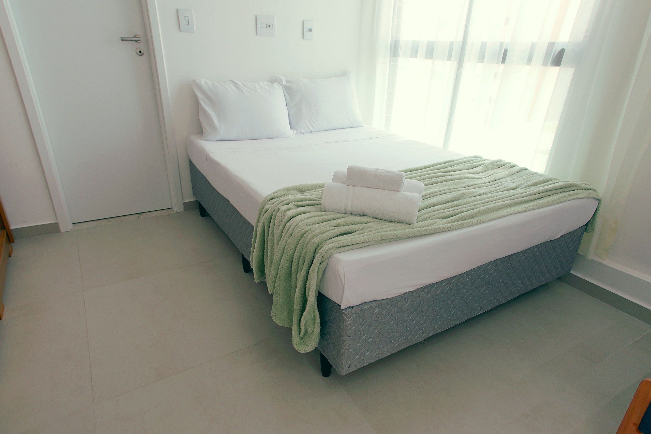 A single bed with a grey headboard, white bedding, and a green throw blanket. There are two white pillows and a folded white towel on the bed. The room has white walls, a white door, and a window with sheer curtains.