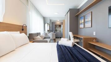 Quarto de hotel com cama, escrivaninha, cadeira, sofá e uma kitchenette ao fundo.