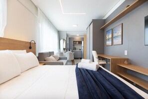 Habitación de hotel con cama, escritorio, silla, sofá y una kitchenette al fondo.