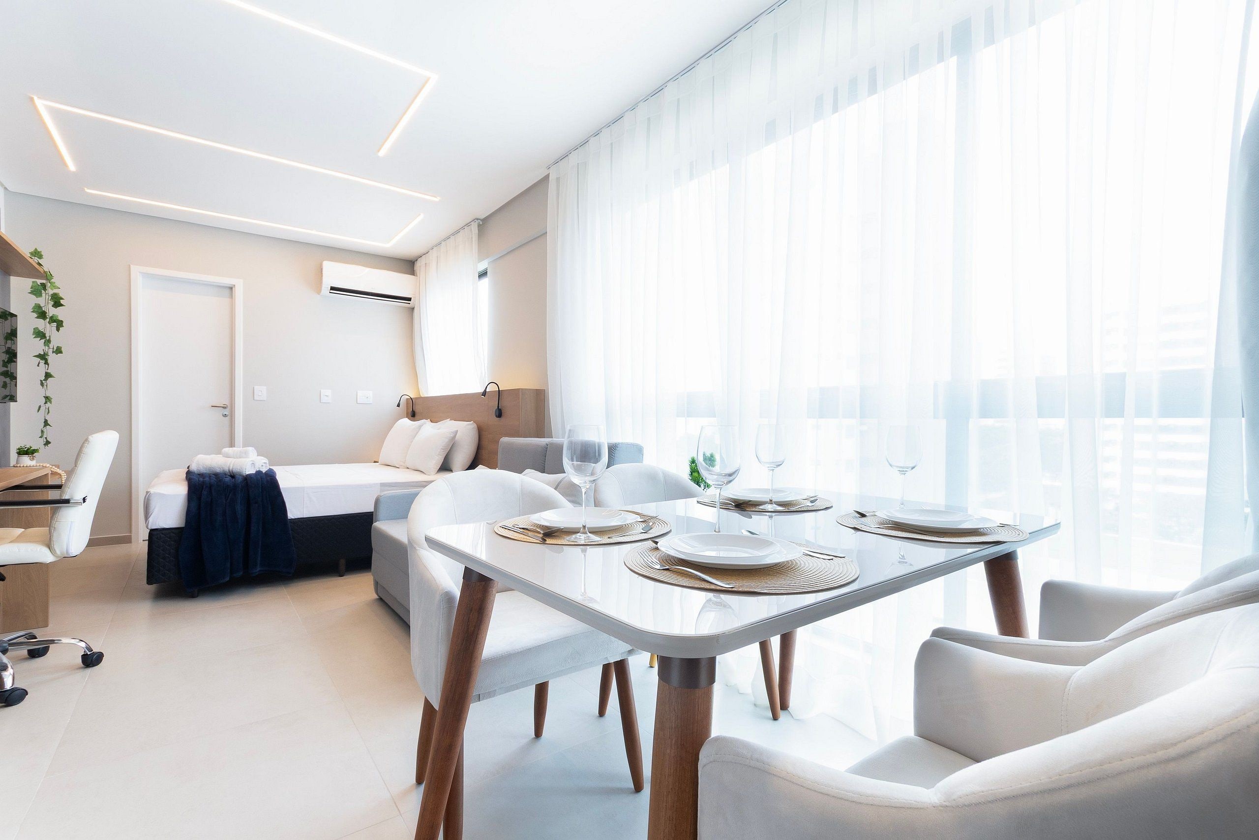 A modern hotel room with a dining area, a bed, and a large window with sheer curtains.