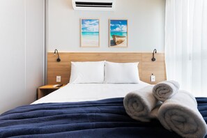 Hotellihuone, jossa on sänky, kaksi seinään kiinnitettyä valaisinta, puinen pääty, yöpöytä, jossa on puhelin, ja kaksi kehystettyä kuvaa seinällä.
