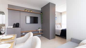 Una habitación de hotel moderna con televisión de pantalla plana, un sofá, una cama, una zona de comedor con mesa y sillas, y una pequeña cocina con cafetera.