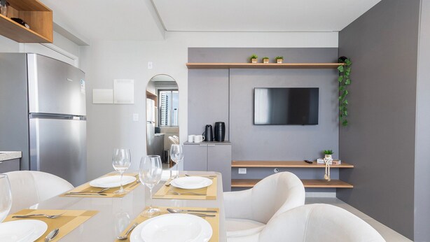 A modern dining area with a stainless steel refrigerator, a flat-screen TV mounted on the wall, and a minimalist design with white walls and wooden shelves. - Botanik With Coworking, Pet Place, and Skyview By Carpediem (Recife)
