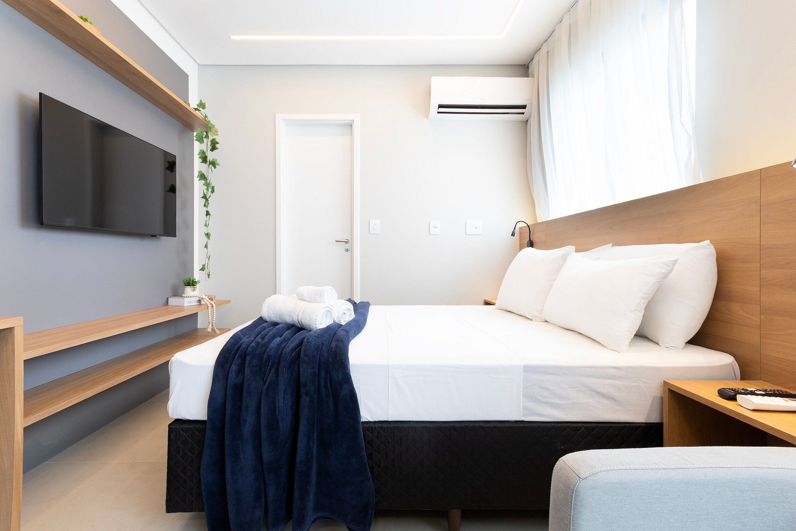 Una habitación de hotel moderna con televisión de pantalla plana, cama con ropa blanca, manta azul, mesita de noche con un control remoto y cabecero de madera.