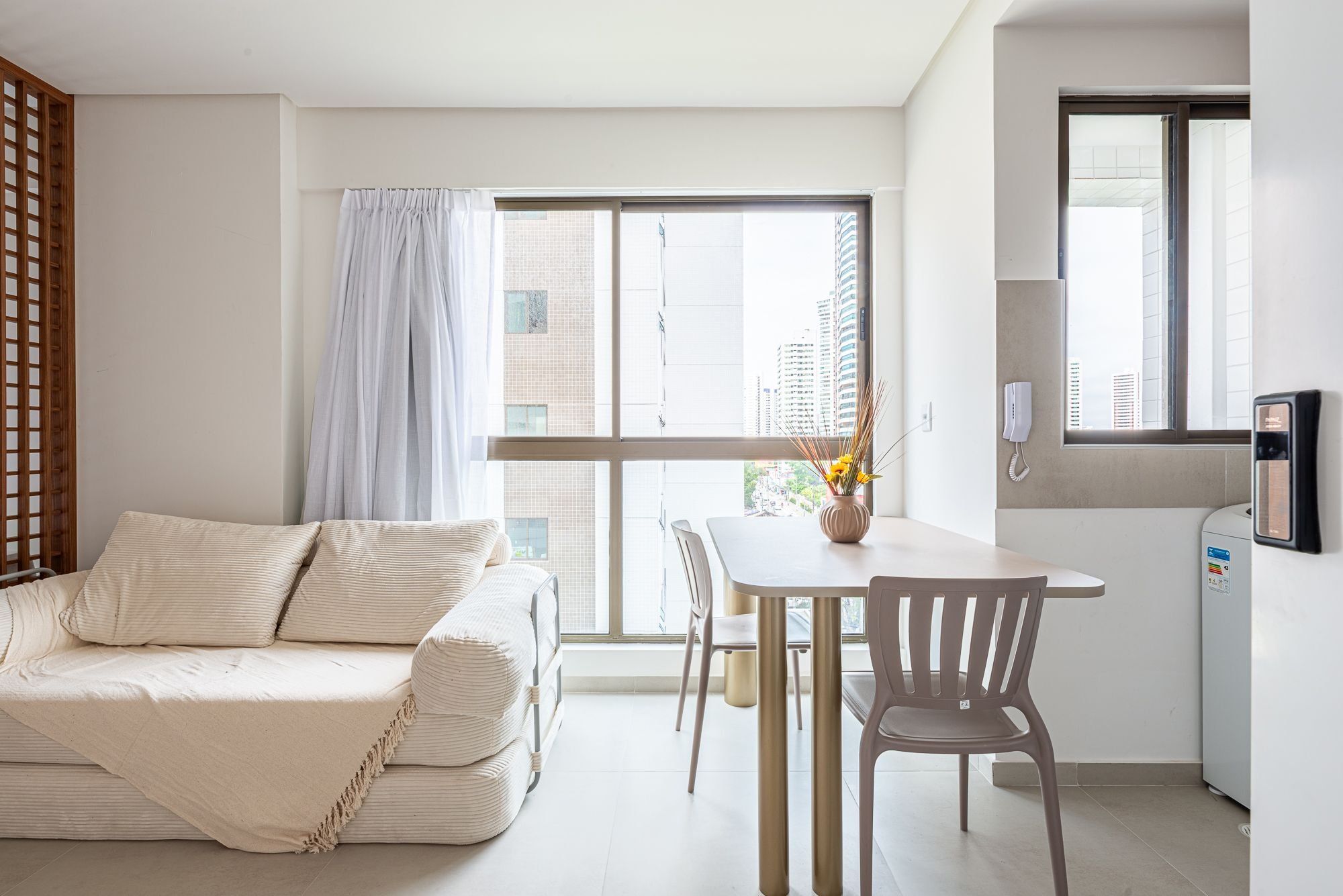 A modern apartment with a sofa, dining table, chairs, a small kitchen area, and large windows offering a city view.