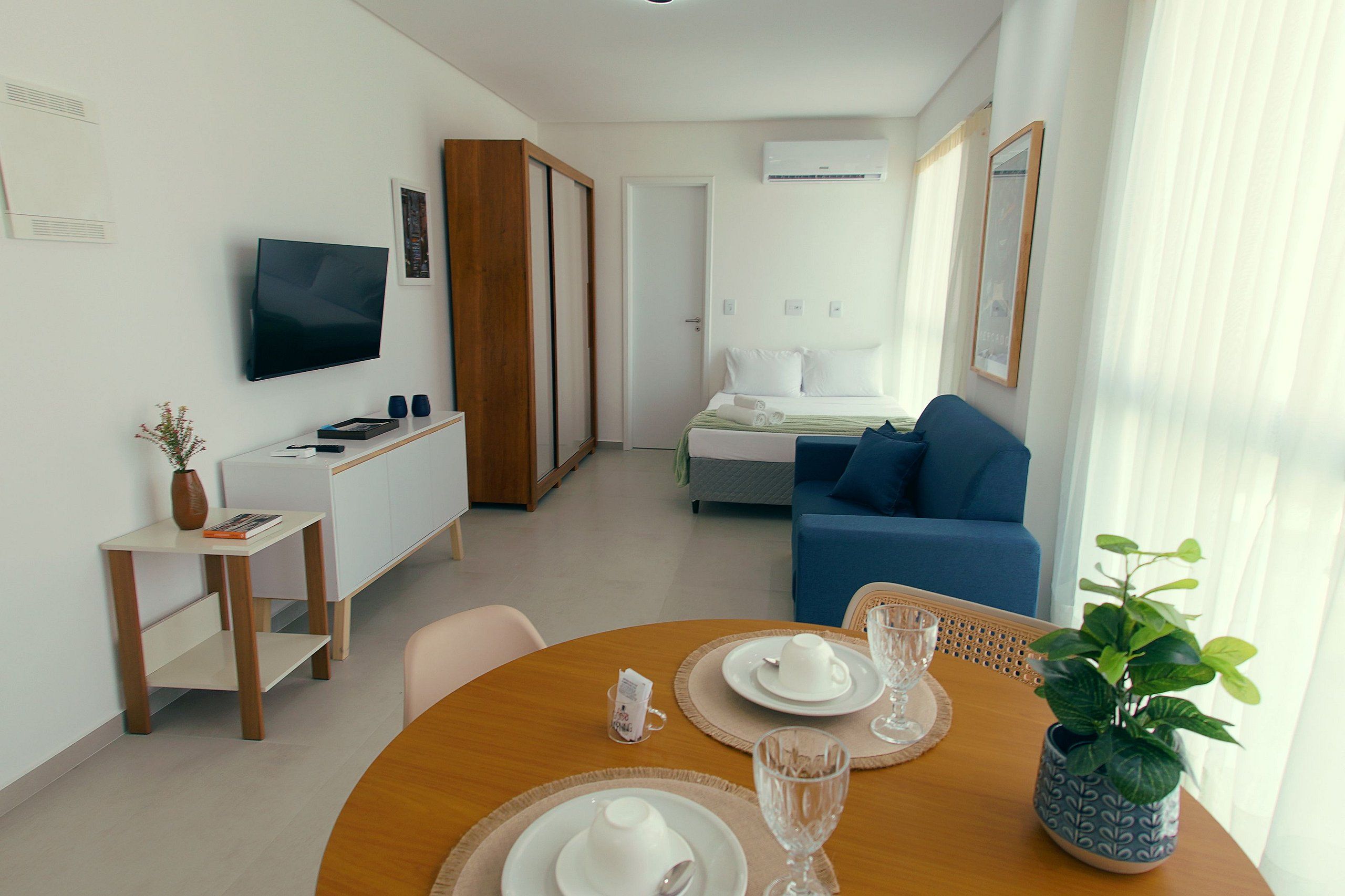 Een hotelkamer met een televisie, een bed, een blauwe bank, een eettafel gedekt met borden en glazen, en een potplant.