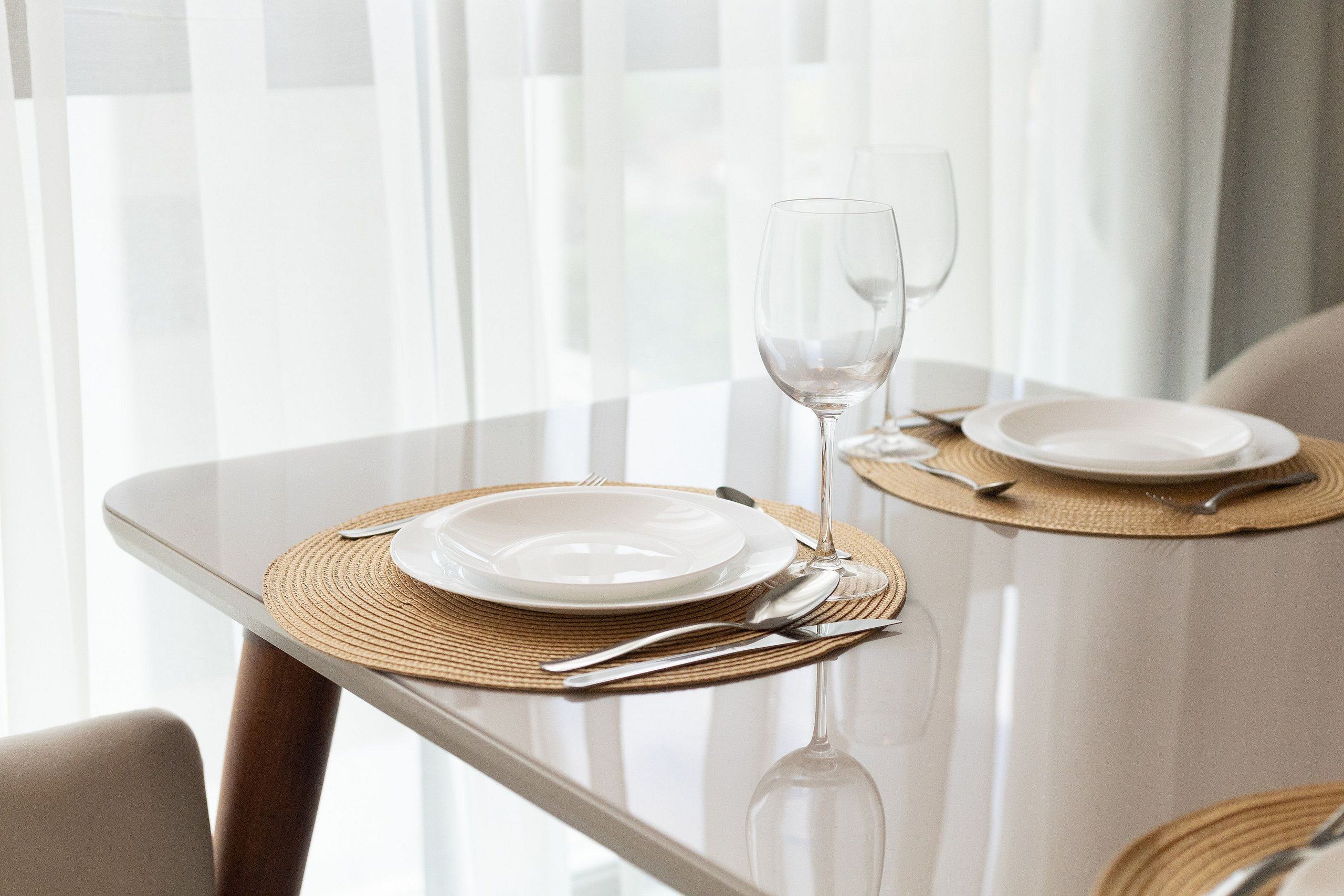 Una mesa de comedor con cubierta de vidrio, dos platos blancos, una copa de vino y cubiertos, todo sobre un mantel individual tejido.