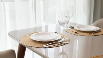 Una mesa de comedor con cubierta de vidrio, dos platos blancos, una copa de vino y cubiertos, todo sobre un mantel individual tejido.