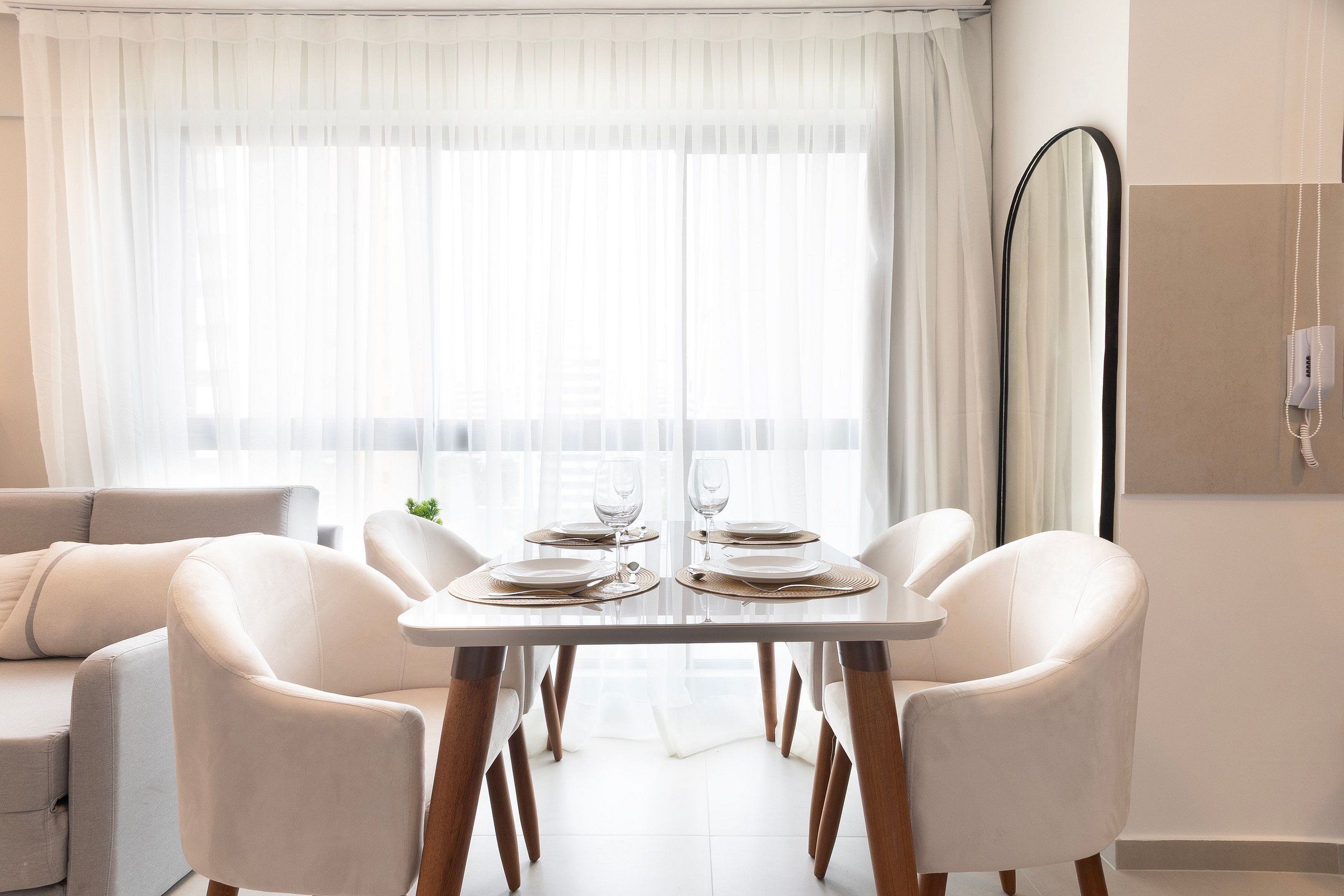 A dining area with a table set for two, white chairs, a sofa, and a large window with sheer curtains.