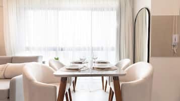 A dining area with a table set for two, white chairs, a sofa, and a large window with sheer curtains.