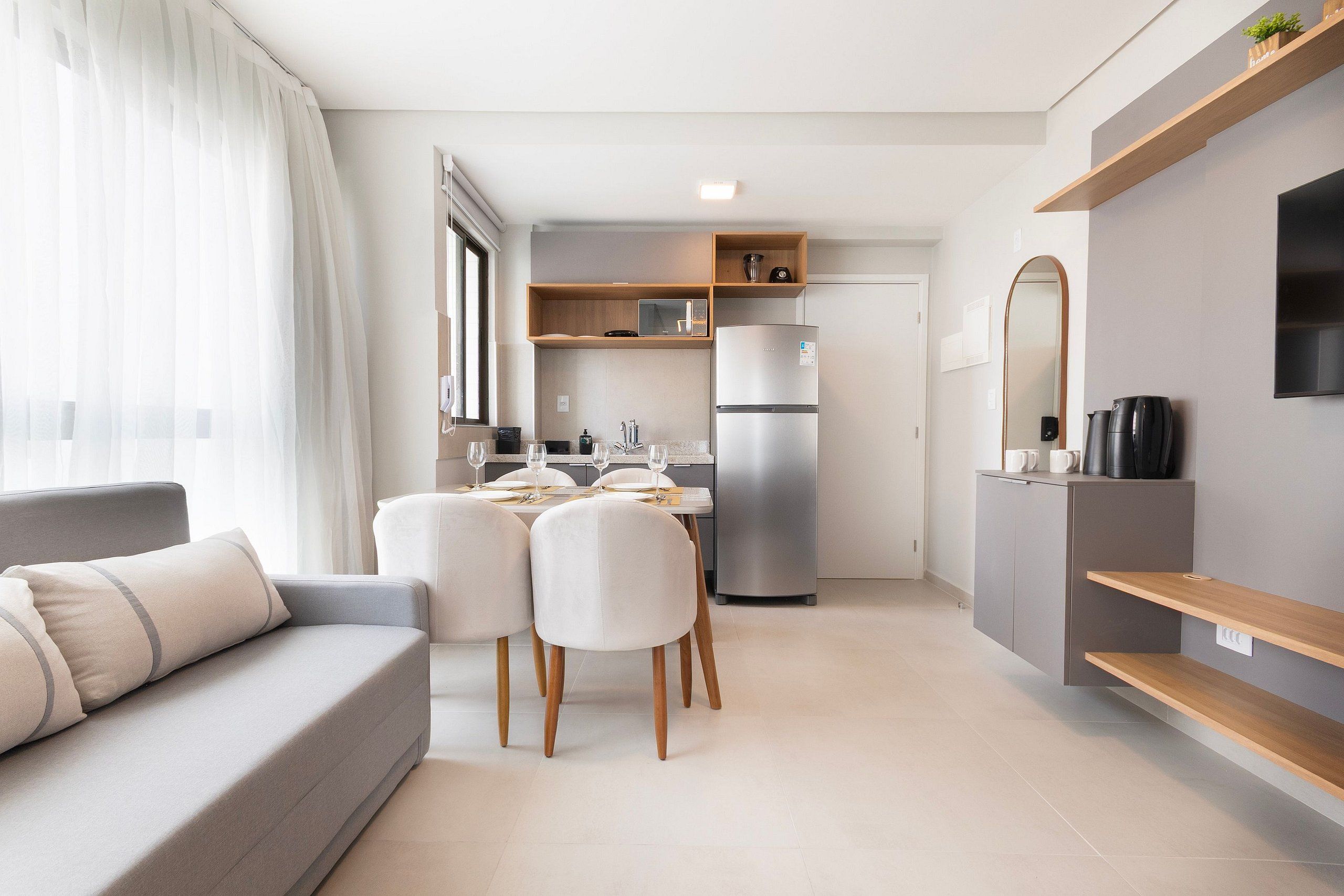 A modern living room with a sofa, dining table, chairs, a kitchen area with a refrigerator, and a mounted TV.