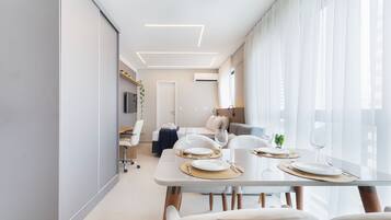 Uma sala de jantar moderna com mesa de vidro, cadeiras brancas e um sofá ao fundo.