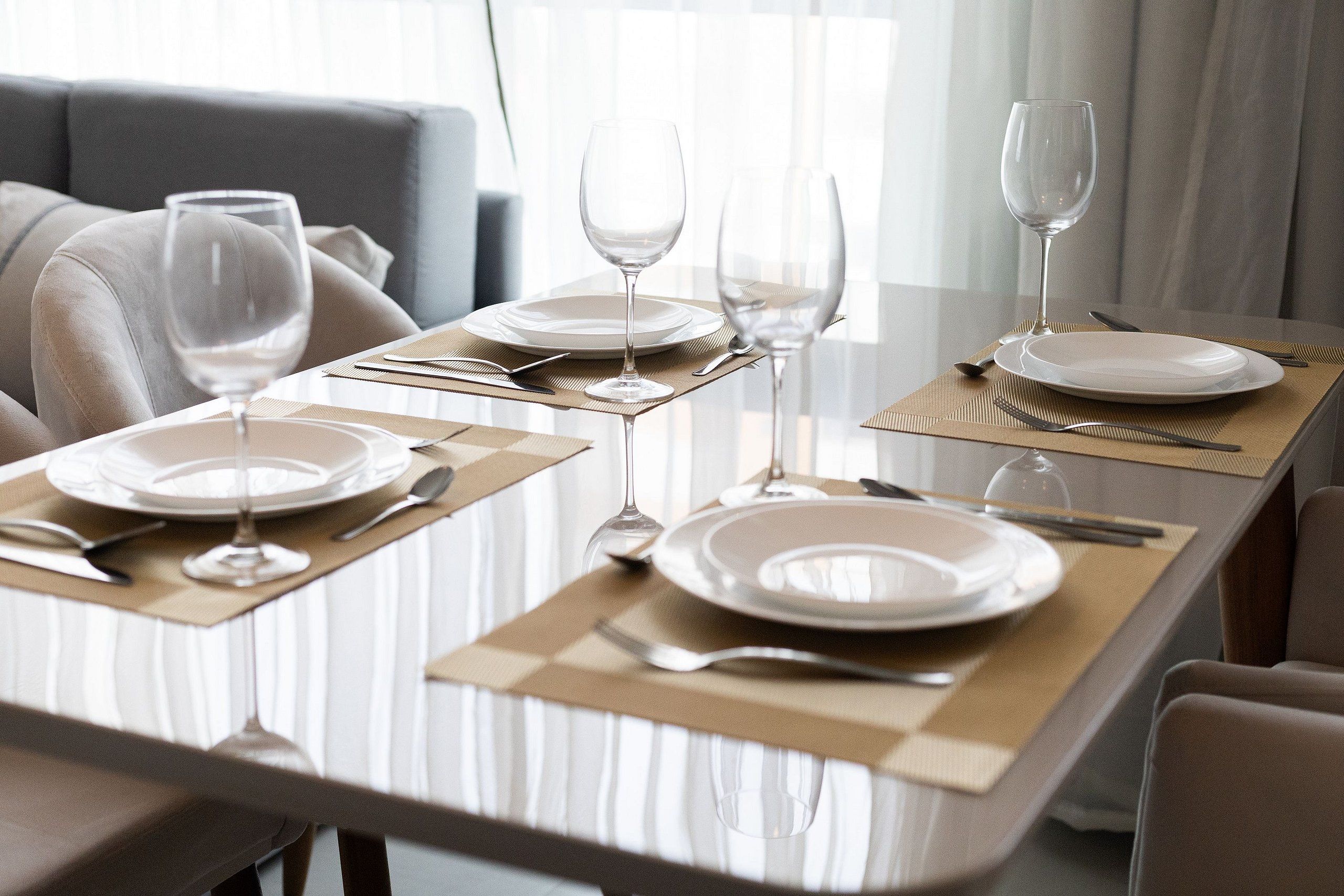 Een eettafel gedekt met borden, bestek en wijnglazen, met een bankstel op de achtergrond.