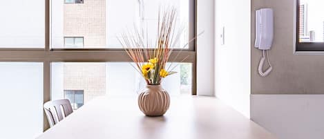A modern dining area with a round table, two chairs, a vase with yellow flowers, and a view of a cityscape through large windows.