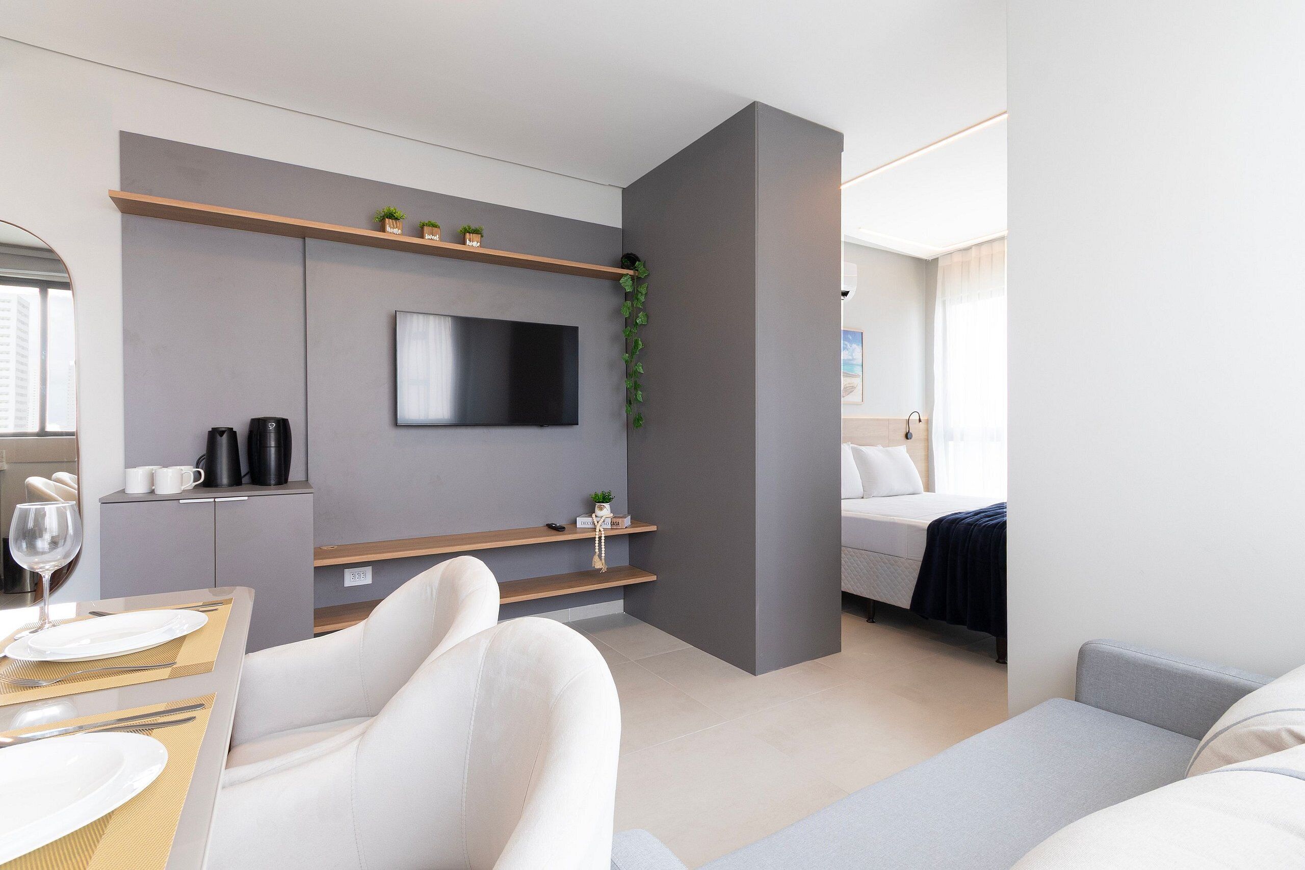 Una habitación de hotel moderna con televisión de pantalla plana, un sofá, una cama, una zona de comedor con mesa y sillas, y una pequeña cocina con cafetera.