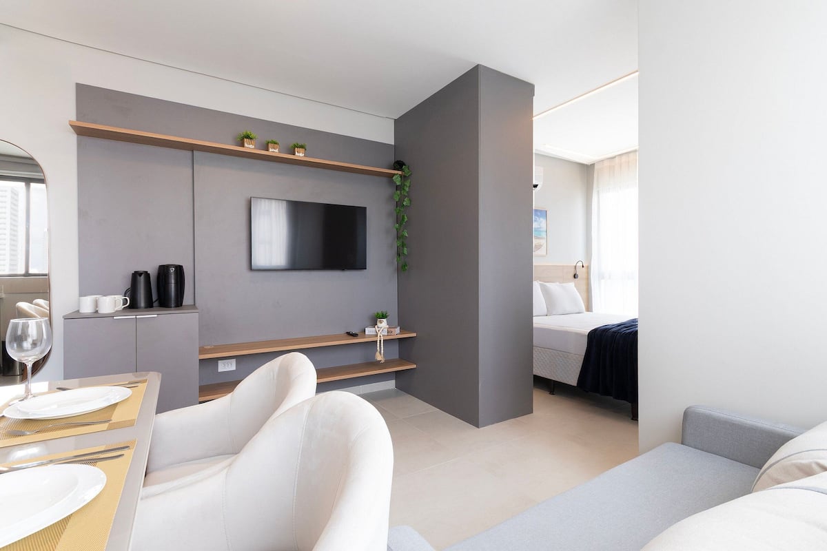 Una habitación de hotel moderna con televisión de pantalla plana, un sofá, una cama, una zona de comedor con mesa y sillas, y una pequeña cocina con cafetera.
