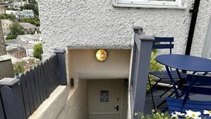 Una entrada de casa con puerta, ventanal pequeño, planta en maceta, y una mesa y sillas azules.