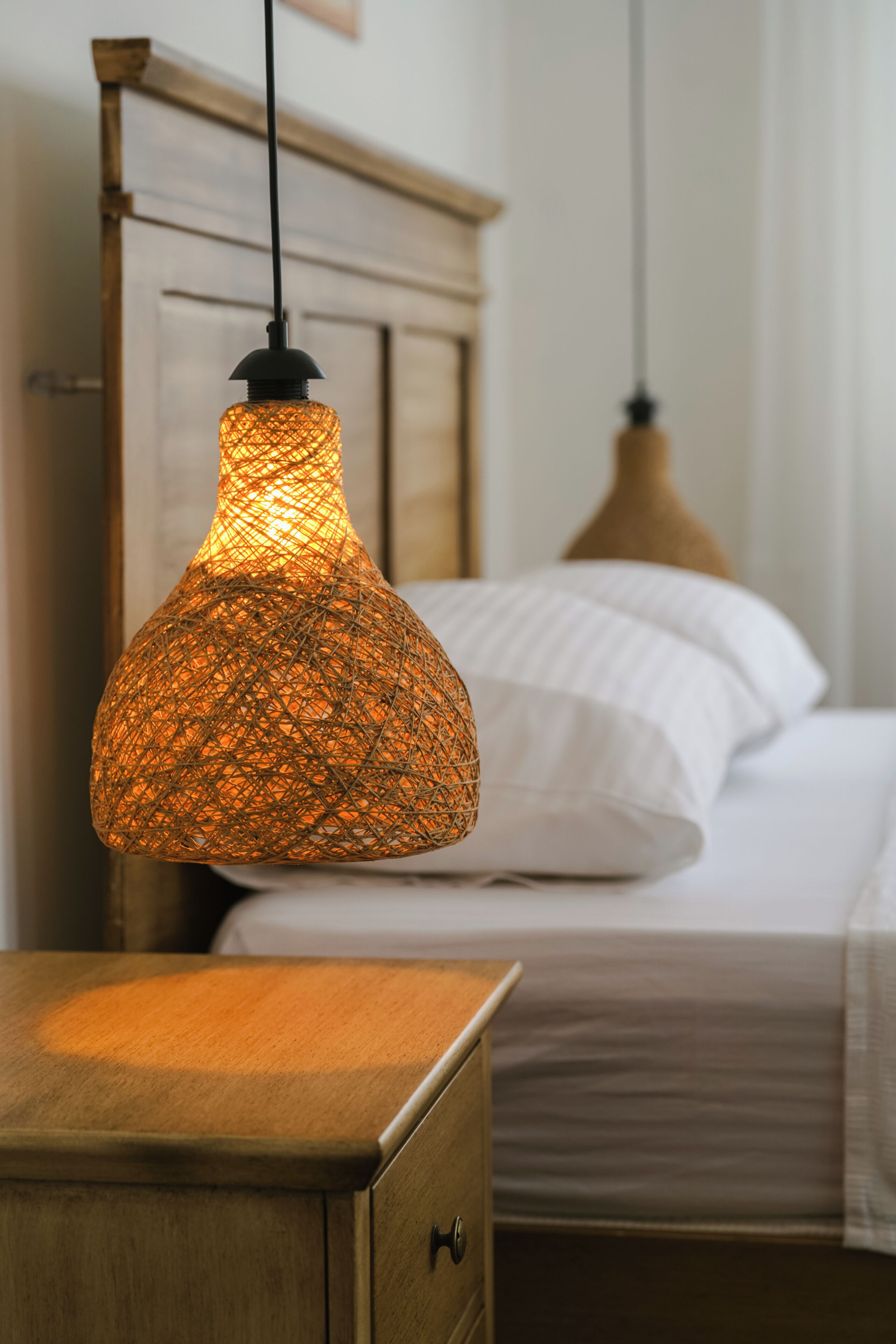 A bed with white linens, a wooden headboard, and a hanging woven light fixture.