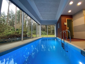 Una piscina interior amb parets de vidre transparent, una escala metàl·lica i vistes a una zona boscosa.