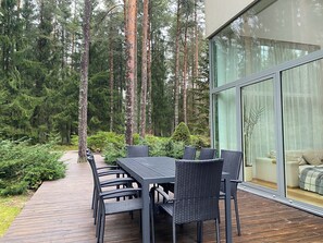 A modern outdoor dining area with a table and chairs, surrounded by a wooden deck and dense forest.