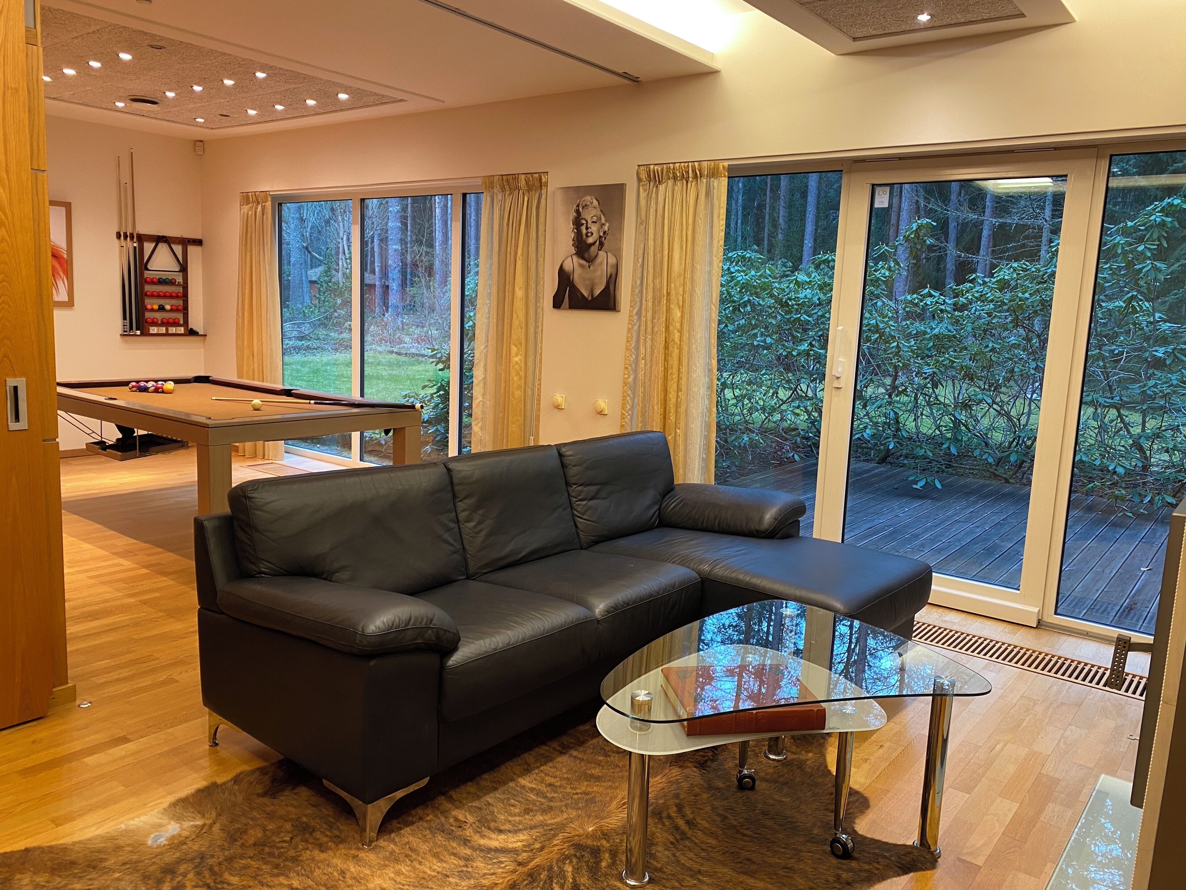 A modern living room with a leather sofa, a glass coffee table, a pool table, and large windows with a view of greenery.