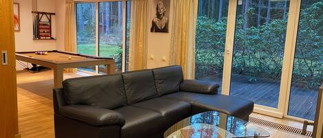 A modern living room with a leather sofa, a glass coffee table, a pool table, and large windows with a view of greenery.