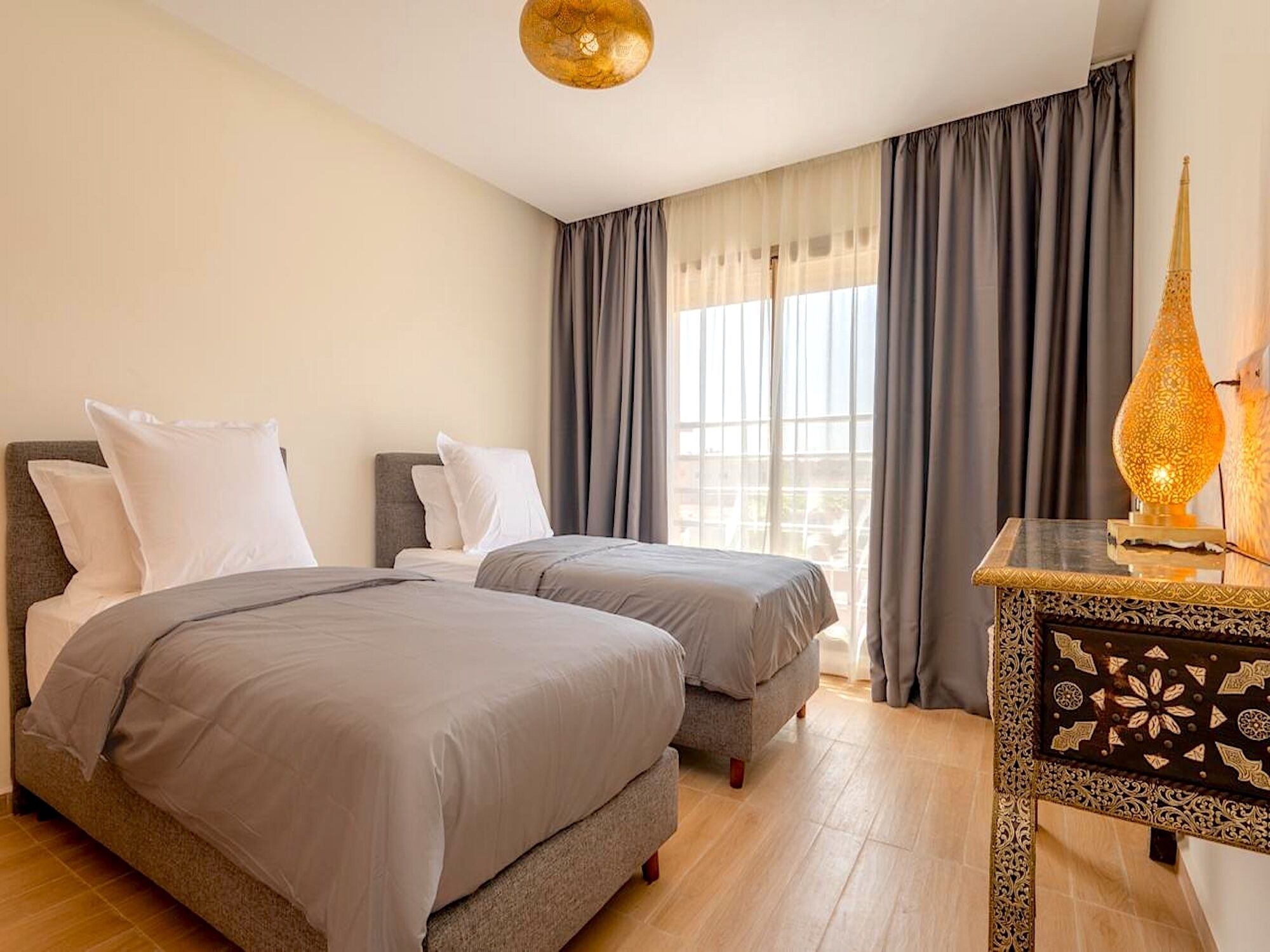 A hotel room with two beds, a nightstand with a lamp, and a view of the city through large windows.