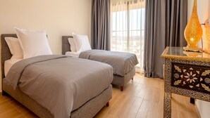 Ein Hotelzimmer mit zwei Betten, einem Nachttisch mit Lampe und Blick auf die Stadt durch große Fenster.