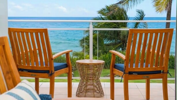 Un balcone con due sedie di legno e un tavolo in vimini, con vista su un mare azzurro e palme.
