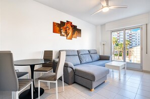 Een moderne woonkamer met een grijze hoekbank, een ronde eettafel met stoelen, een plafondventilator en een groot raam met uitzicht op gebouwen.
