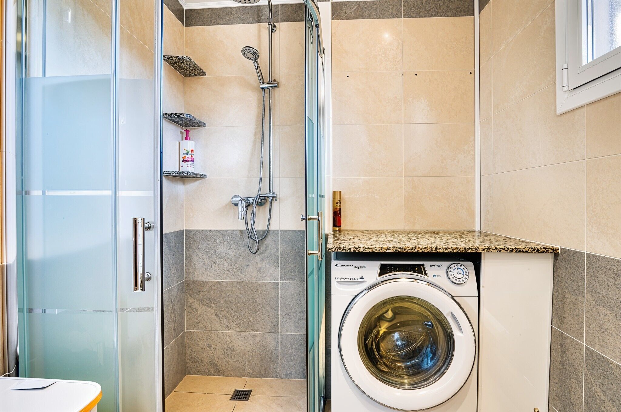 Een compacte badkamer met een glazen douchecabine, een ingebouwde wasmachine en een betegelde vloer.