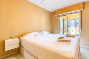 Een slaapkamer met een bed, een nachtkastje, een raam met uitzicht op gebouwen en een houten plafondpaneel.