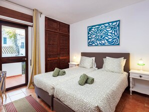 Ein Schlafzimmer mit einem Bett, zwei Nachttischen, einem Fenster mit Vorhängen, einer Tür und einem an der Wand hängenden abstrakten Kunstwerk.