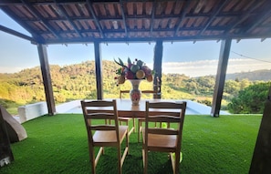 Un patio techado con una mesa y sillas, un florero con fruta, y vistas a una ladera boscosa.