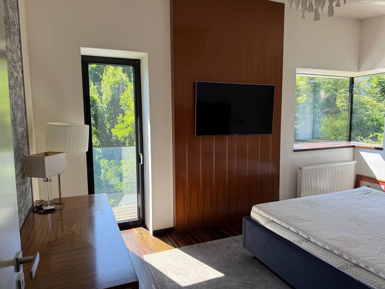 Ein modernes Schlafzimmer mit Bett, einem an der Wand befestigten Fernseher, einem Schreibtisch mit Lampe und Blick auf Grünflächen durch das Fenster.