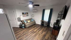 Un espace de vie compact comprenant un coin cuisine, un canapé agrémenté de coussins décoratifs, une table à manger avec des chaises et un ventilateur de plafond.