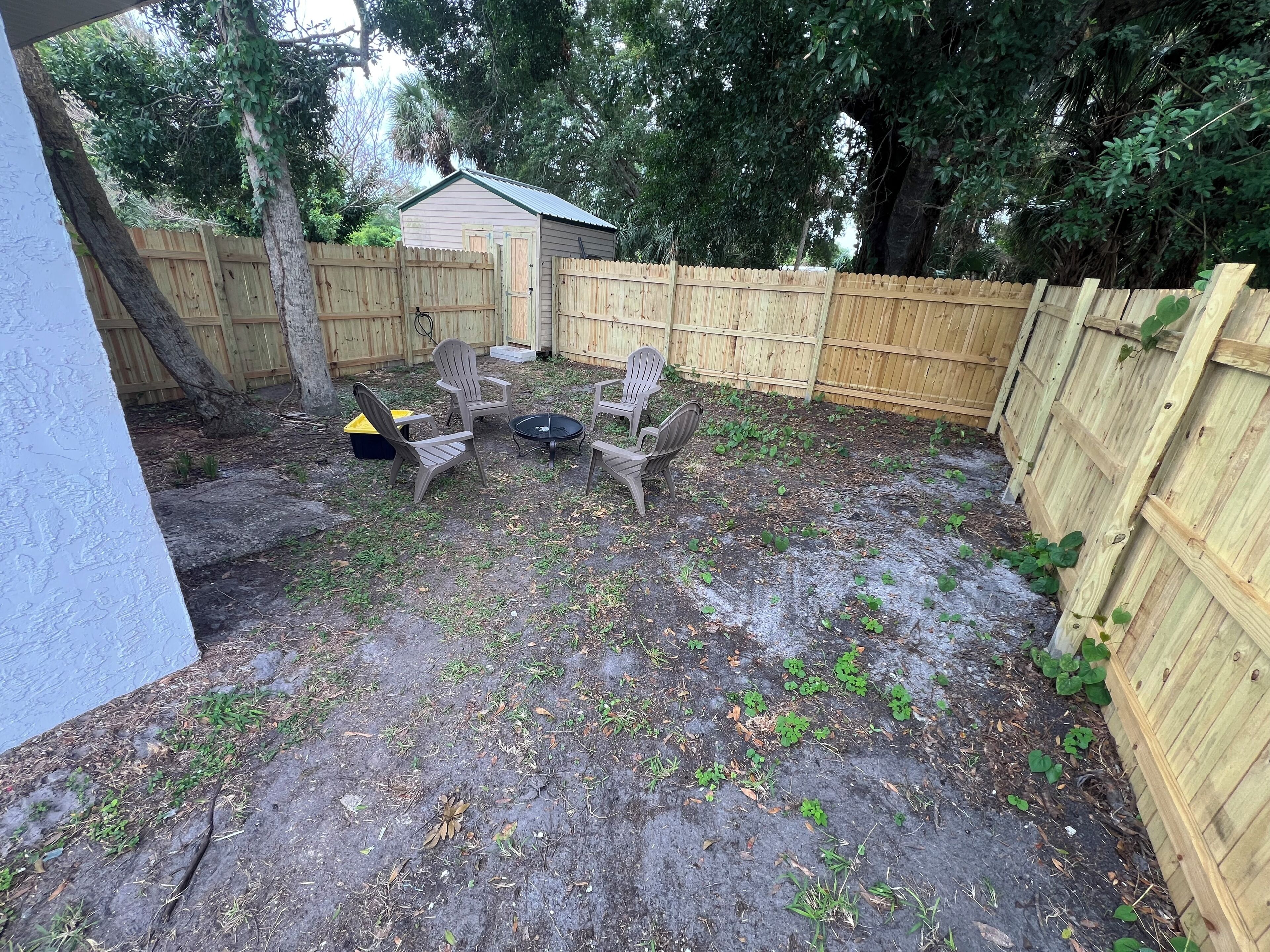 A backyard with a wooden fence, a small shed, and a patio area with chairs and a table.