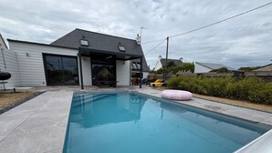 Ein rechteckiger Pool mit einer Terrassenanlage, einem überdachten Sitzbereich und einem Haus im Hintergrund.