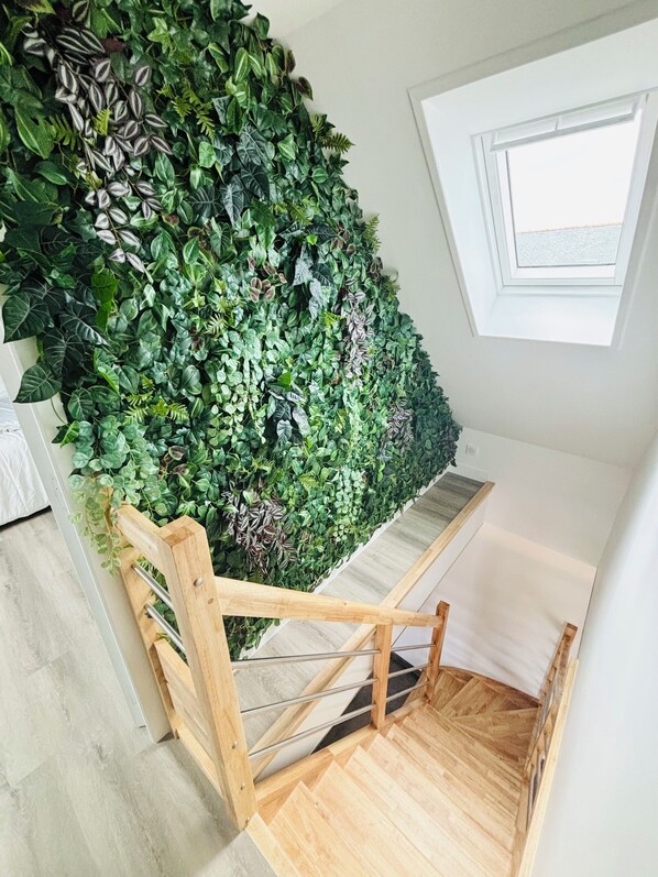 A staircase with a wooden handrail, a green wall with various plants, and a window with a view of the sky.
