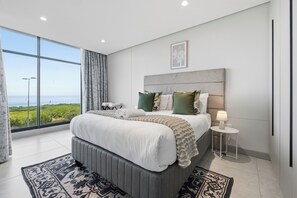 Ein modernes Schlafzimmer mit einem großen Bett, einem Nachttisch mit Lampe und Blick ins Freie.