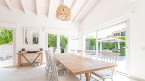 Uma sala de jantar ampla com uma mesa de madeira e cadeiras brancas, janelas grandes e um ventilador de teto.